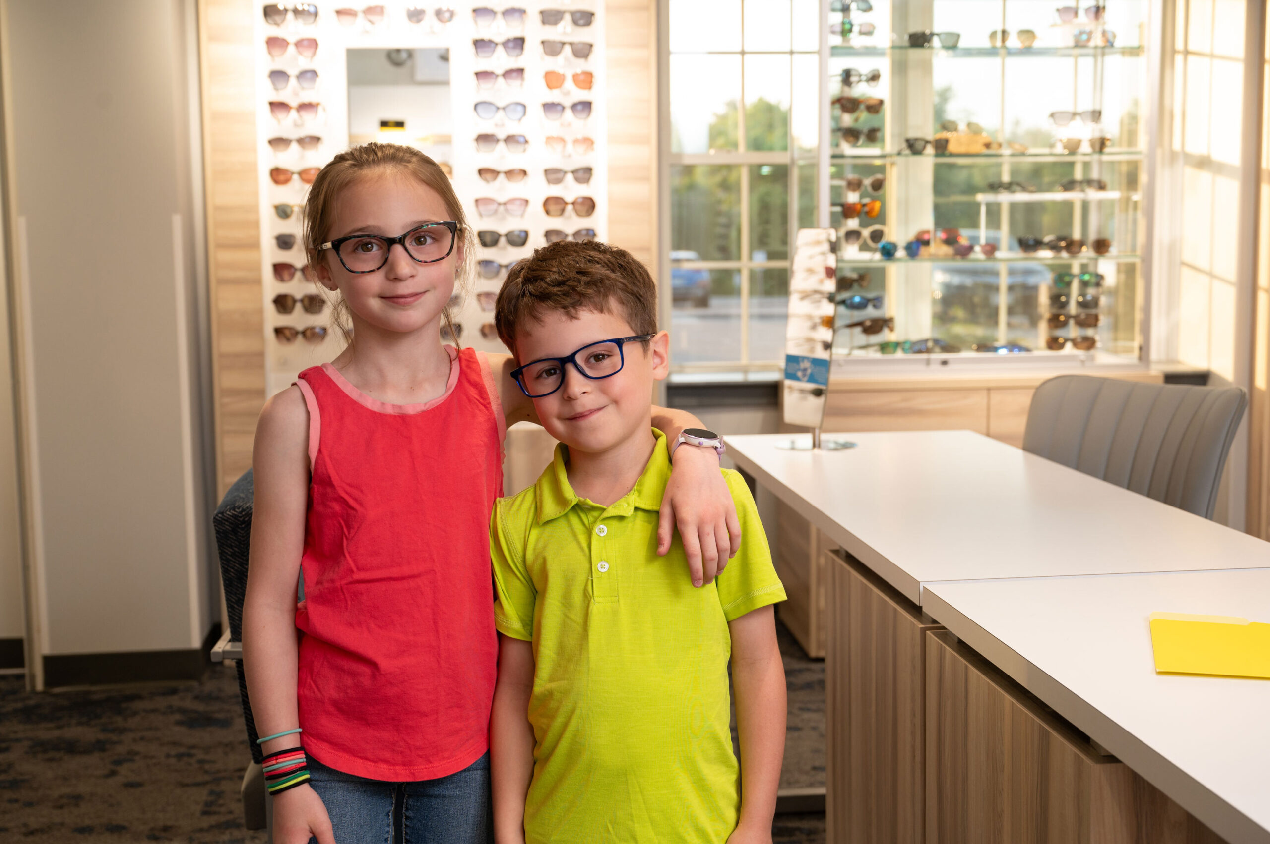 Two children wearing glasses stand together and smile inside an optical store with display shelves behind them.