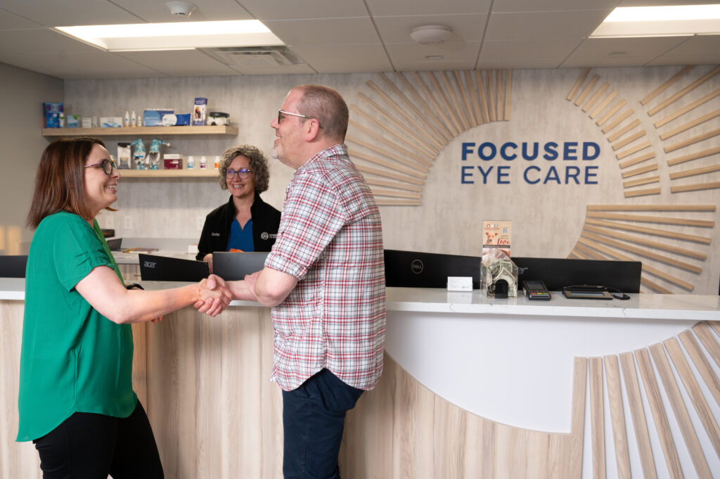 Two people shake hands at an eye care clinic reception while a staff member smiles behind the counter.