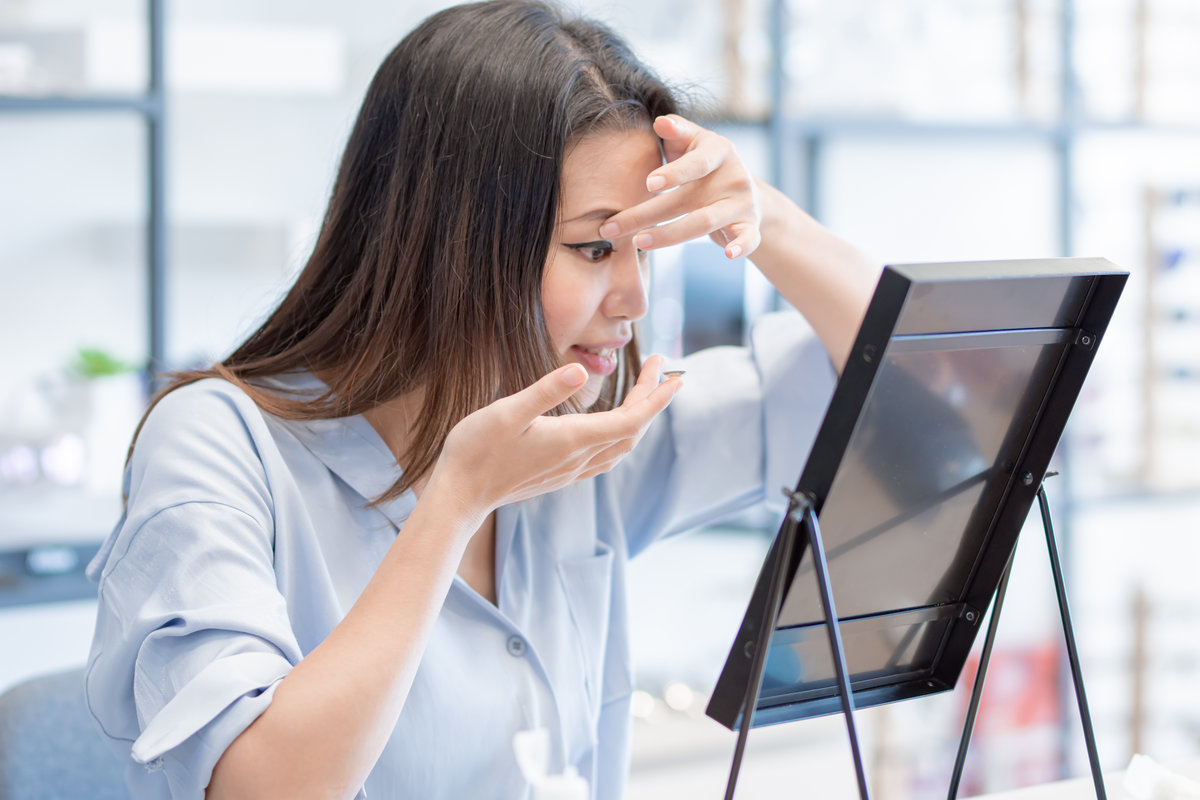 Woman putting in a contact lens while looking into a mirror on a table in a bright room.