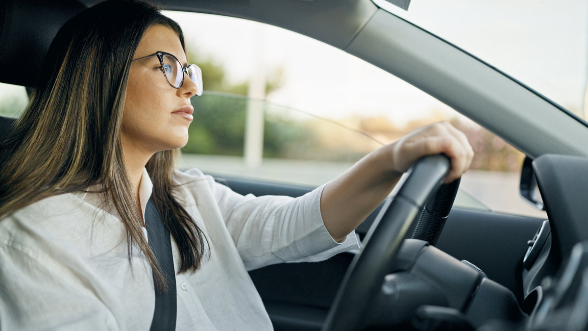 Woman with glasses driving a car, looking thoughtful and focused while holding the steering wheel.