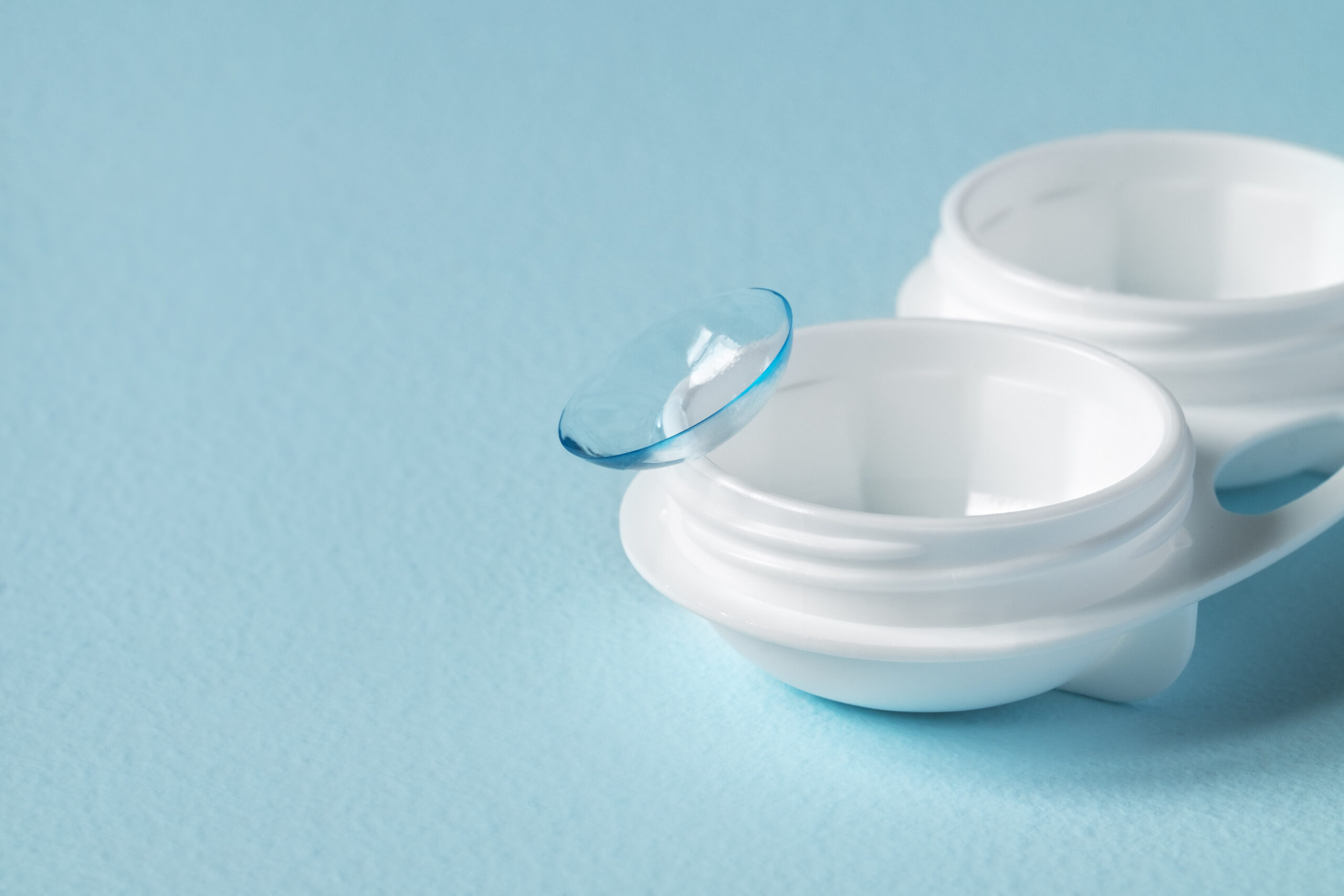 A contact lens rests on an open white contact lens case against a light blue background.