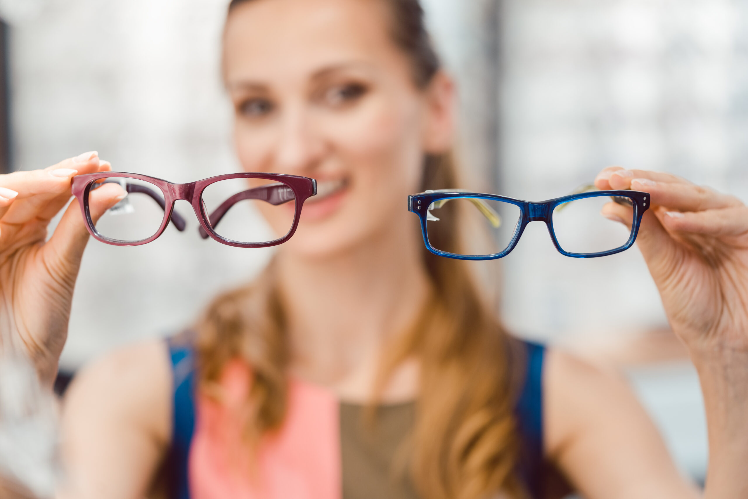 Smiling woman holding up two pairs of eyeglasses, one purple and one blue, for comparison.
