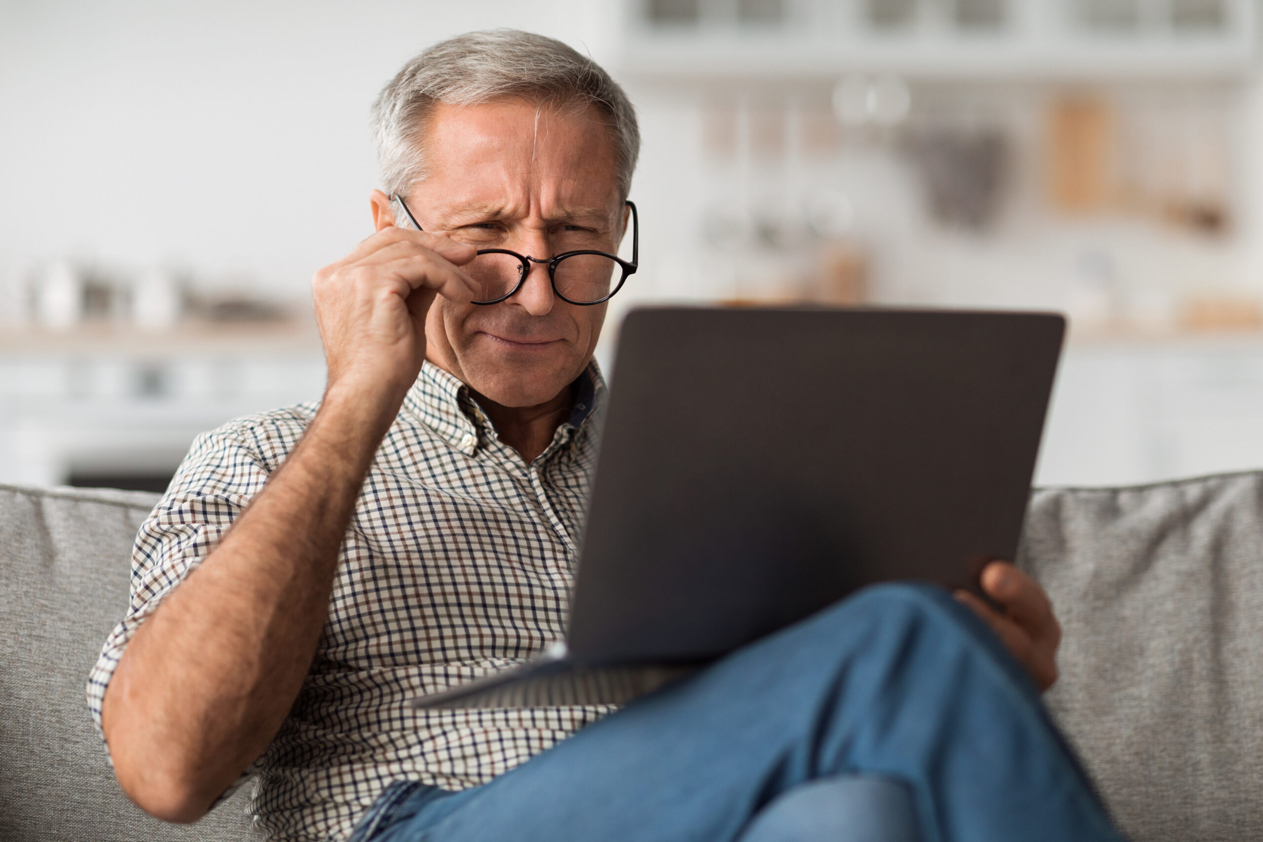 Older man in glasses squinting at a laptop screen while sitting on a sofa, looking confused or focused.