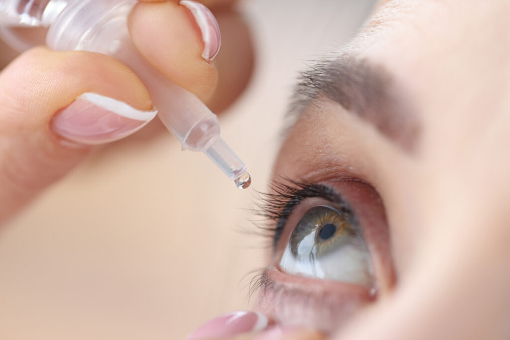 Close-up of a person applying eye drops to their right eye with a hand holding the dropper.