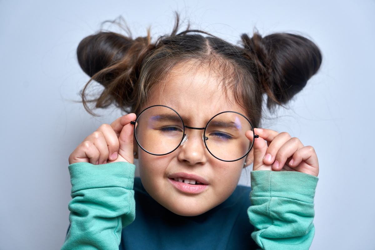 Young girl with double buns squints and holds large round glasses, wearing a blue-green sweatshirt.