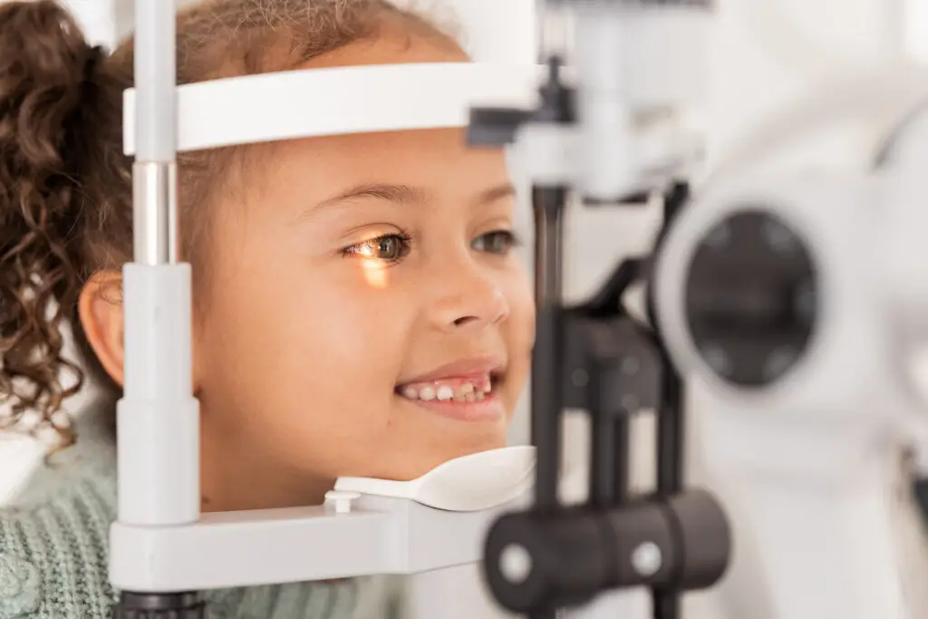 Smiling child getting an eye exam with a slit lamp at an optometrists office.