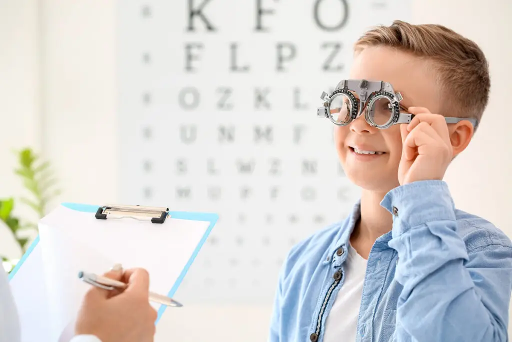 Smiling boy wearing trial frames during an eye exam, with an eye chart and clipboard in view.
