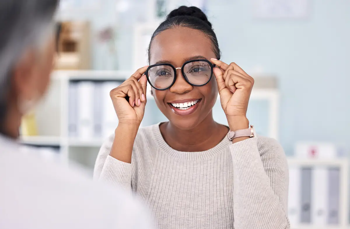 Smiling woman trying on eyeglasses in an office setting, facing another person out of focus in the foreground.