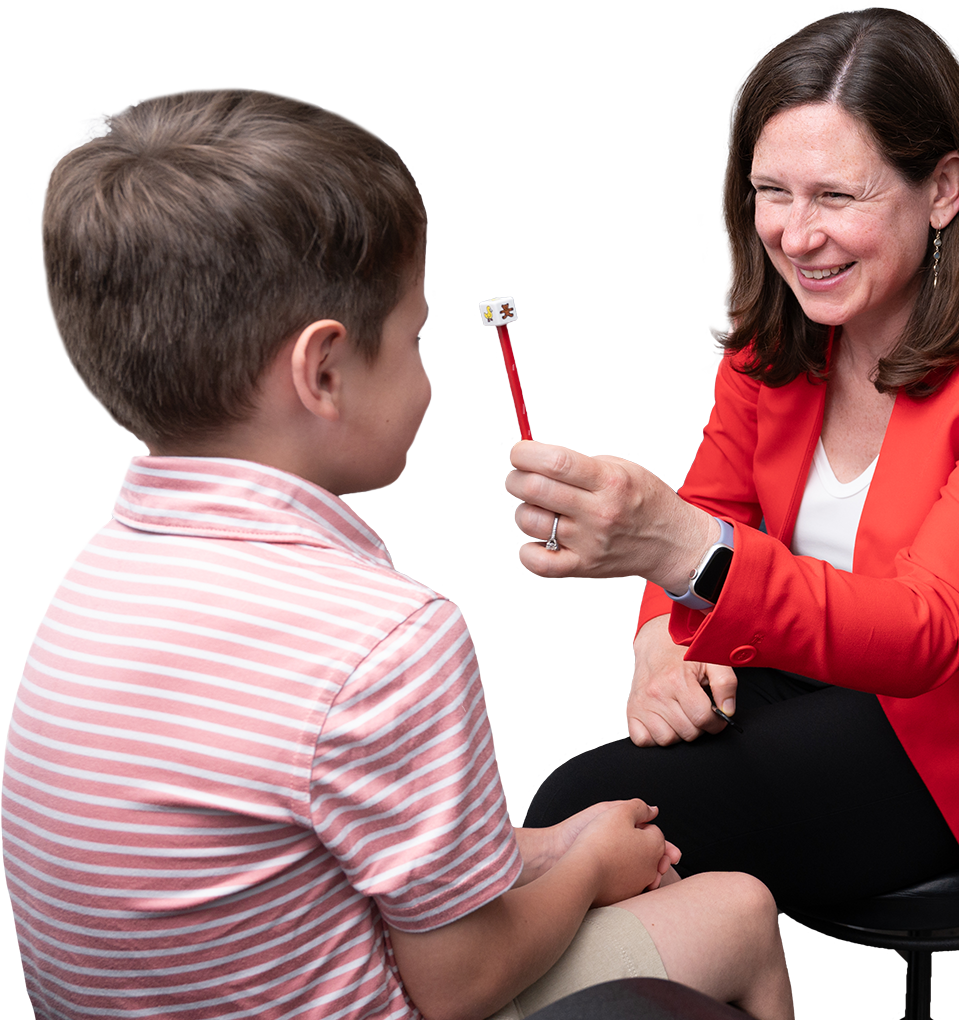 A woman in a red blazer holds a pencil in front of a seated boy wearing a pink striped shirt.