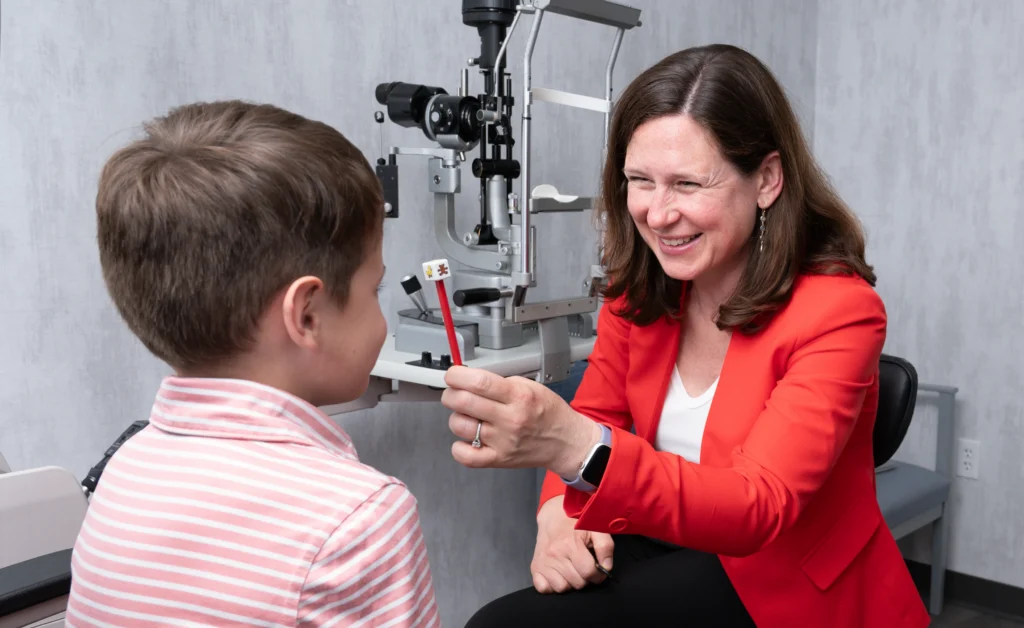 Woman in red jacket holds up an object, smiling at a boy during an eye exam in a clinic.