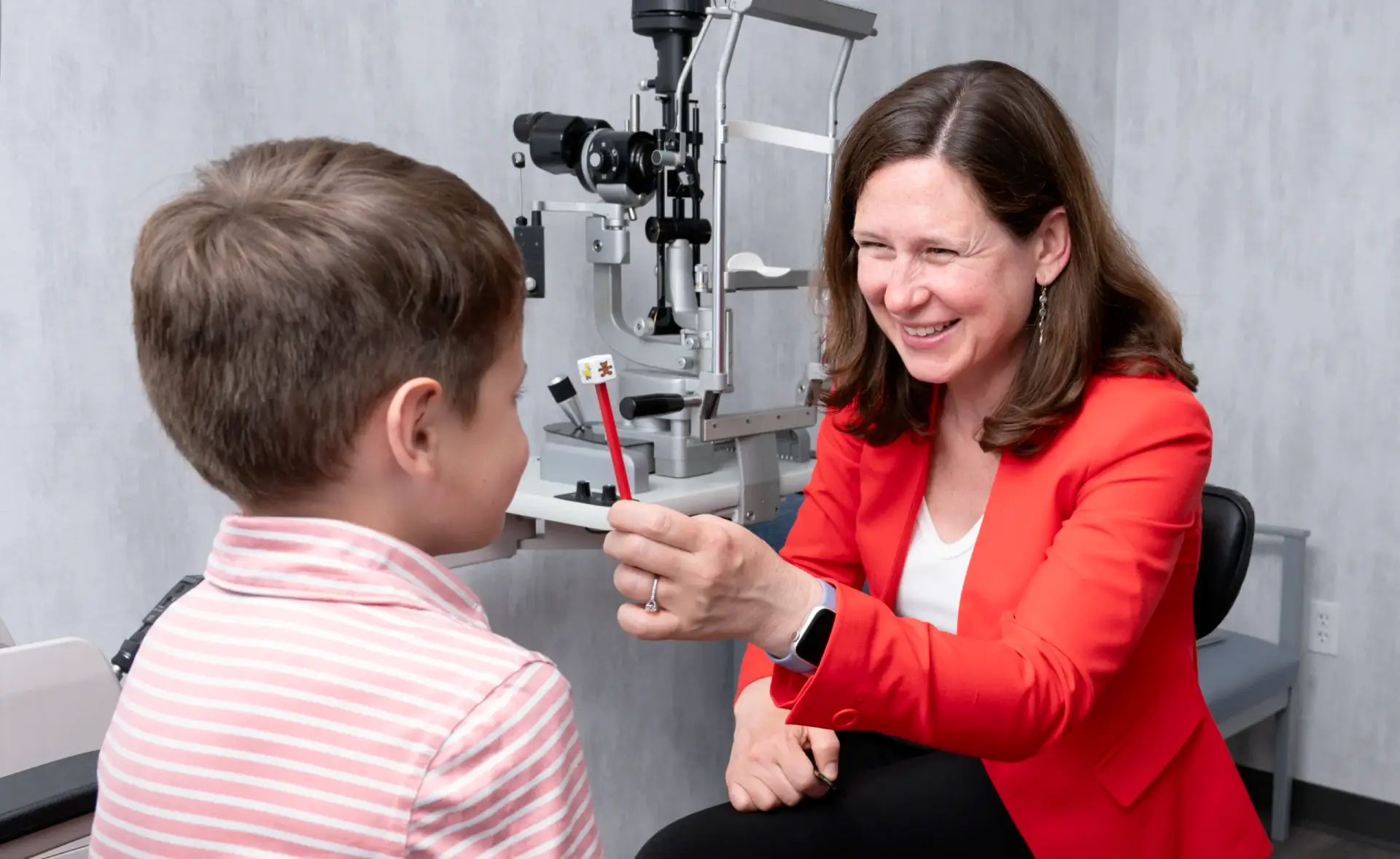 Woman in red jacket holds up an object, smiling at a boy during an eye exam in a clinic.
